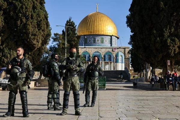 Group of soldiers in tactical gear standing in a stone courtyard with the Golden Dome of the Rock in Jerusalem in the background.
