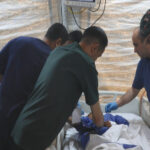 Medical team performing an emergency procedure at a hospital bed inside a tent-like field setup, with gloves and masks on.