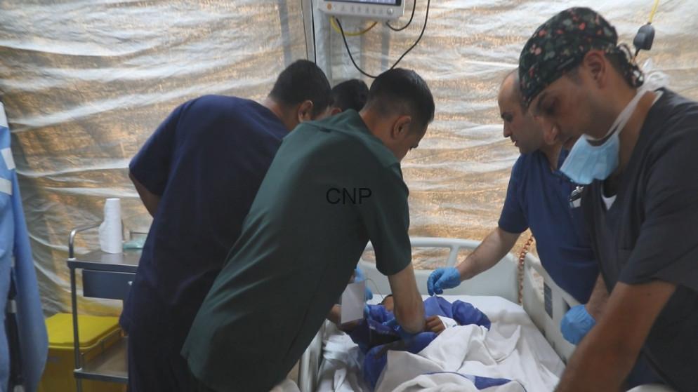 Medical team performing an emergency procedure at a hospital bed inside a tent-like field setup, with gloves and masks on.