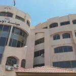 Exterior view of a beige multi-story building with curved glass facade and Arabic sign, under a blue sky (Media Commission building)