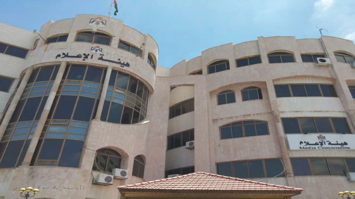 Exterior view of a beige multi-story building with curved glass facade and Arabic sign, under a blue sky (Media Commission building)