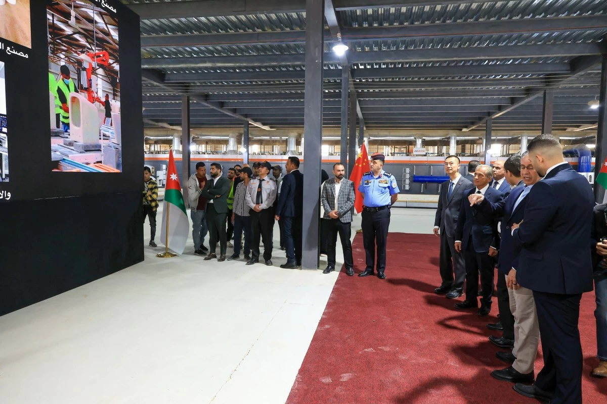 Group of officials and workers listening to a presentation inside a factory, with Jordan and China flags visible on stands to the left and center.