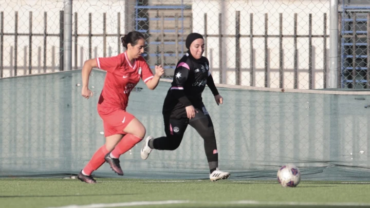 - Center News Network Two female soccer players chase a ball on an outdoor field; one in a red kit, the other in black with a hijab, near the fence and goal area.