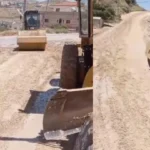 Two construction vehicles on a dusty rural road: a road roller in the foreground and a wheel-loader behind it, leveling the dirt road.