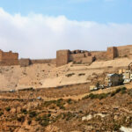 Ancient stone fortress wall and ruins perched on a sandy hillside above a small town of beige houses.