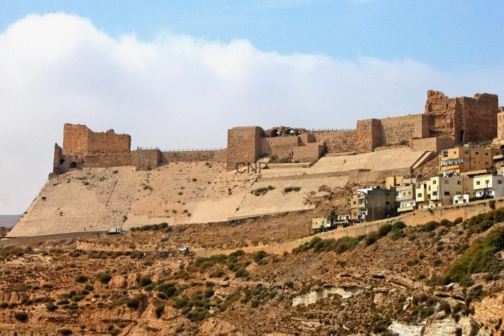 - Center News Network Ancient stone fortress wall and ruins perched on a sandy hillside above a small town of beige houses.