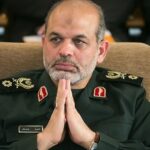 Middle-aged man in a dark green military uniform sits with hands pressed together in a prayer-like gesture, a serious expression on his face.