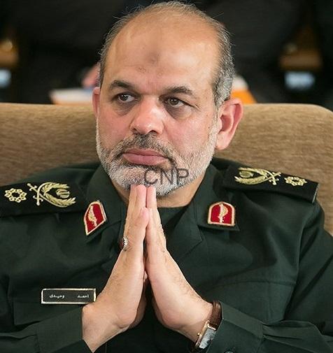 Middle-aged man in a dark green military uniform sits with hands pressed together in a prayer-like gesture, a serious expression on his face.