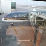 Small private jet parked at a terminal gate, connected to a jet bridge, with water visible beyond the tarmac.