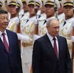 Two men in dark suits walk side by side past a row of ceremonial guards in white uniforms and gold sashes.
