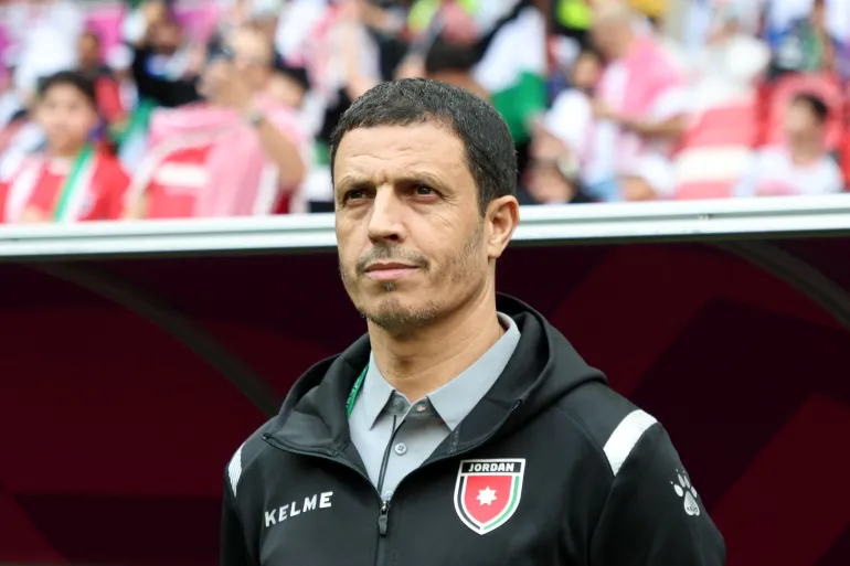 Athletic coach standing on a sideline, wearing a black Kelme jacket with a Jordan crest, with a blurred stadium crowd in the background.