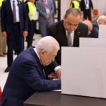 Man in a dark suit signing a ballot at a polling booth, with another man overseeing nearby