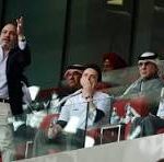 Group of people standing on a glass-front balcony, some raising hands and cheering in a crowded venue.