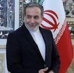 Man in a dark suit standing indoors beside the Iranian flag, smiling, with ornate wall decor in the background.