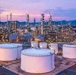 Oil refinery at dusk with large white storage tanks, pipes and processing towers, and a distant city skyline.