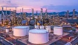 Oil refinery at dusk with large white storage tanks, pipes and processing towers, and a distant city skyline.