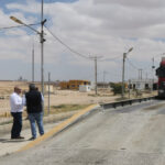 Two men stand on a rural road beside a raised weigh bridge as a red truck crosses; a police officer watches on the right.