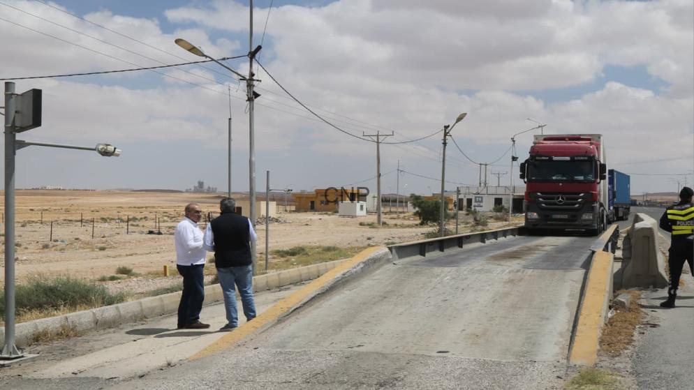 ash - Center News Network Two men stand on a rural road beside a raised weigh bridge as a red truck crosses; a police officer watches on the right.