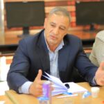 Man in a navy blazer and light blue striped shirt seated at a table, speaking and gesturing with papers on the desk.