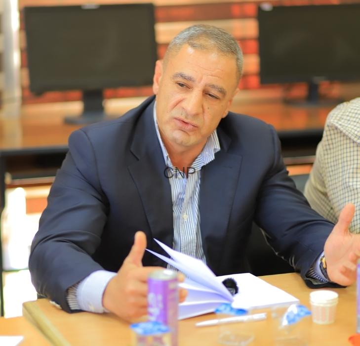 ayman - Center News Network Man in a navy blazer and light blue striped shirt seated at a table, speaking and gesturing with papers on the desk.