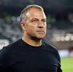 A middle-aged man in a black athletic shirt on a football pitch, looking ahead with a stadium in the background.