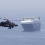 Military helicopter hovering near a large cargo ship at sea, as if preparing to deck or land or inspect the vessel (CNP watermark visible).
