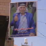 Campaign banner showing a man in a blue suit giving a thumbs-up, mounted on a street with Arabic caption at the bottom.