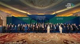 Large group photo of conference attendees in formal attire on a stage in a grand hall, with a blue banner behind them.