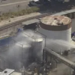 Industrial facility with a cylindrical storage tank and steam venting near rail tracks.
