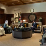 Diplomatic meeting in a wood-paneled room with officials seated in two groups and a Jordan flag nearby, a clock on the wall.