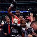 Boxer in the ring raising a championship belt in victory, with reporters and photographers around him.
