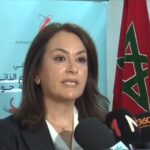 Woman in a blazer speaks into microphones at a press conference, with a Moroccan flag on the right and a blue banner behind her.