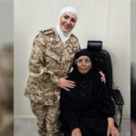 Female soldier in desert camouflage uniform and white hijab stands beside an elderly woman in black seated in a chair, both smiling indoors.