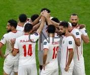 Group of football players in white jerseys celebrate in a huddle on a green pitch, raising their hands together in triumph.