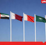 Row of five national flags fluttering on tall flagpoles against a clear blue sky, suggesting an international or diplomatic setting.