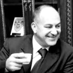 Smiling man in a suit and tie, holding a small cup, in a room with ornate woodwork behind him.