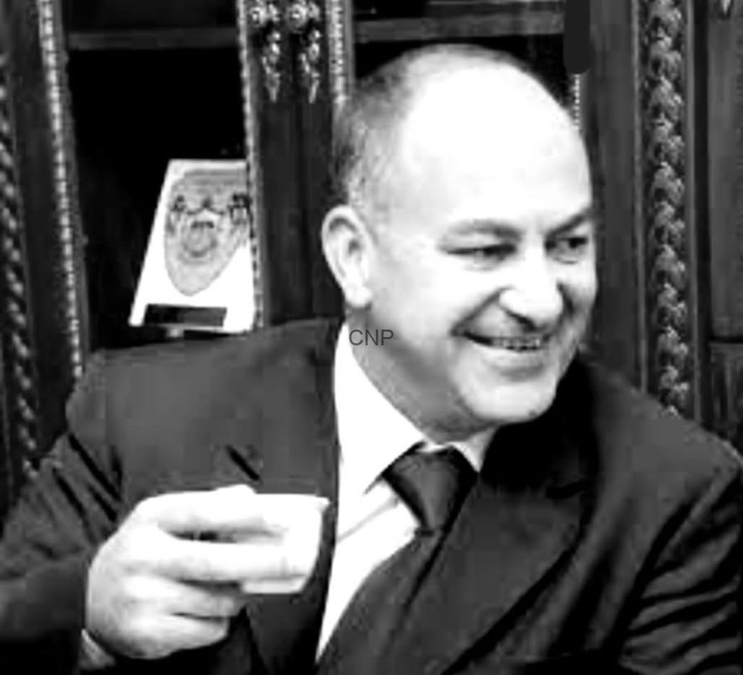 hash - Center News Network Smiling man in a suit and tie, holding a small cup, in a room with ornate woodwork behind him.