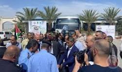 hjj - Center News Network A man in a suit speaks to or greets a crowd at an outdoor event, with a bus and palm trees in the background and photographers nearby.
