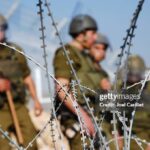 Barbed wire in the foreground with blurred soldiers in combat gear standing behind, suggesting a military checkpoint or border area.