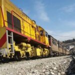 Yellow freight locomotive pulling a line of freight cars on a railroad track in a rural area, with a blue sky and hillside town in the distance.