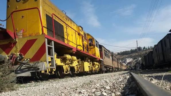 Yellow freight locomotive pulling a line of freight cars on a railroad track in a rural area, with a blue sky and hillside town in the distance.