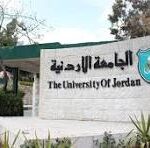 Entrance sign for The University of Jordan with its emblem and surrounding plants and gate in the background.