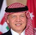 Man wearing a red-and-white checkered keffiyeh and white thobe, smiling with flags in the background.
