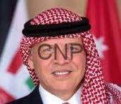 Man wearing a red-and-white checkered keffiyeh and white thobe, smiling with flags in the background.