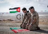 Two soldiers kneeling on a rocky beach, paying respects to a Jordanian flag laid flat on the ground.
