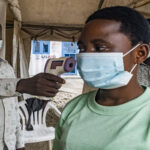 Person in a green shirt with a mask has her temperature checked with a handheld infrared thermometer under a tent, as another masked man looks on.