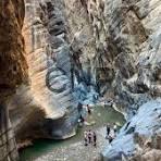 Visitors descend a narrow, rugged canyon with a small turquoise pool at the bottom, flanked by tall, layered sandstone walls.