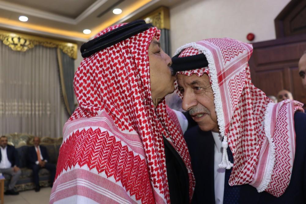 Two men wearing red-and-white keffiyehs greet with a kiss on the forehead in a formal room.