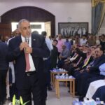 A man in a suit speaks into a microphone at a formal gathering, addressing a seated audience in a large hall.