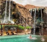 maen - Center News Network Resort with multiple waterfalls cascading into a turquoise pool, lounging area under a thatched canopy, and cliffs in the background.